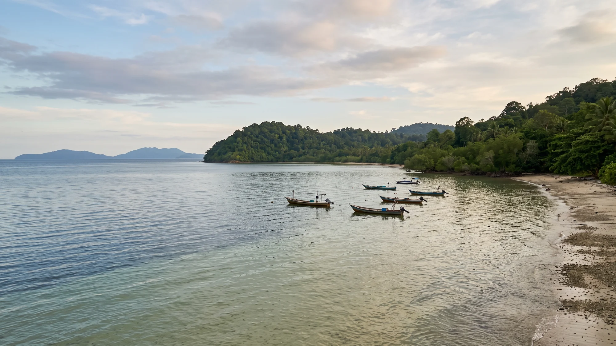 A wide view along a mainland coastline in late afternoon light, shallow water in the foreground and low islands as soft silhouettes on the horizon.