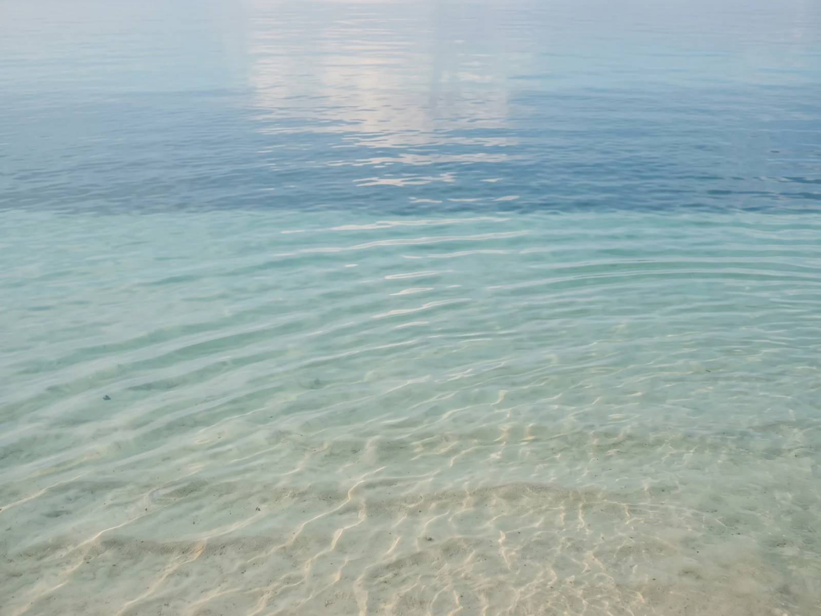 Shallow sea water in low afternoon light — bands of pale turquoise and deeper blue over a sandy bottom.